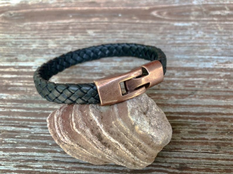 Peut inclure: Un bracelet en cuir tress&eacute; noir avec une fermeture en cuivre. Le bracelet est sur un rocher.