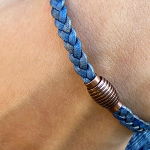 K&ouml;nnte beinhalten: Nahaufnahme eines geflochtenen blauen Lederarmbands mit einem kupferfarbenen Magnetverschluss. Das Armband wird am Handgelenk getragen und zeigt die Textur und Farbe von Leder und Metall.