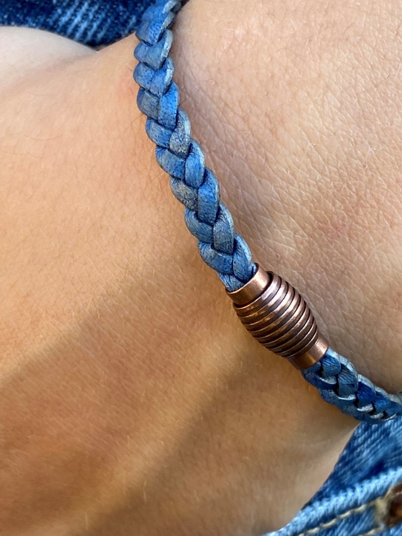 K&ouml;nnte beinhalten: Nahaufnahme eines geflochtenen blauen Lederarmbands mit einem kupferfarbenen Magnetverschluss. Das Armband wird am Handgelenk getragen und zeigt die Textur und Farbe von Leder und Metall.