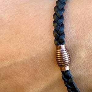 K&ouml;nnte beinhalten: Geflochtenes schwarzes Lederarmband mit einem kupferfarbenen zylindrischen Verschluss. Das Armband wird am Handgelenk getragen, wodurch die Textur und das Design hervorgehoben werden. Ein schlichtes, aber stilvolles Schmuckst&uuml;ck.