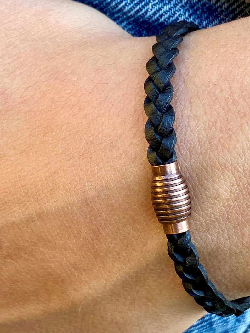 K&ouml;nnte beinhalten: Geflochtenes schwarzes Lederarmband mit einem kupferfarbenen zylindrischen Verschluss. Das Armband wird am Handgelenk getragen, wodurch die Textur und das Design hervorgehoben werden. Ein schlichtes, aber stilvolles Schmuckst&uuml;ck.