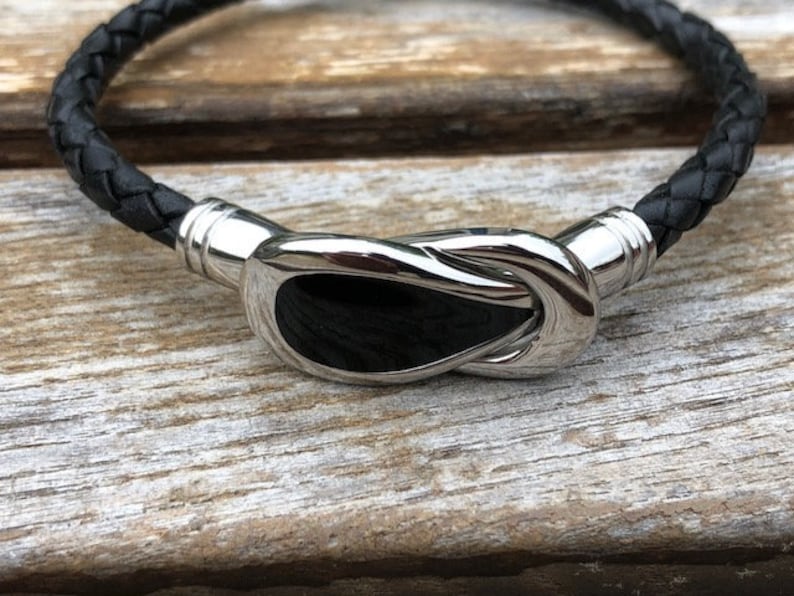 May include: A black leather bracelet with a silver clasp featuring a knot design. The clasp has a black inlay.