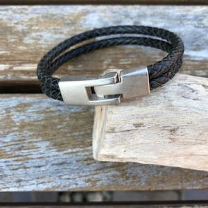May include: A black leather bracelet with a silver clasp. The bracelet is braided and has a simple design.