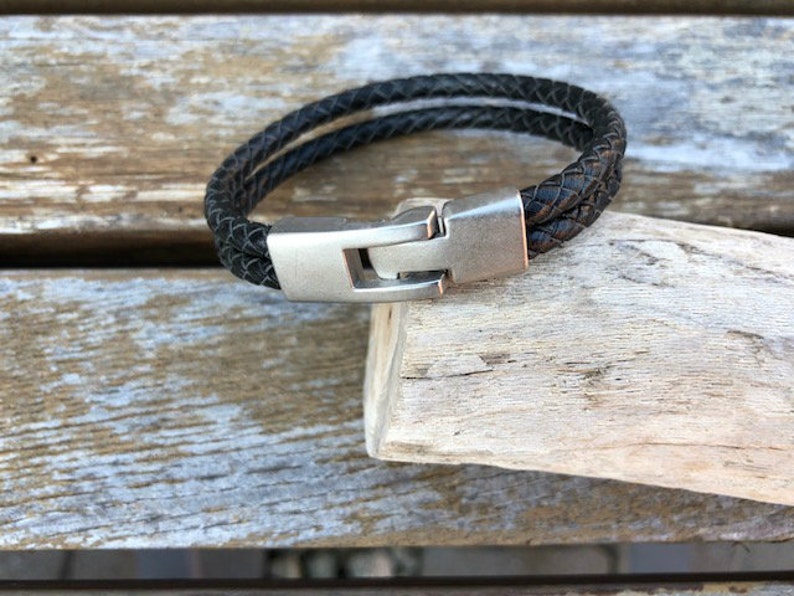 May include: A black leather bracelet with a silver clasp. The bracelet is braided and has a simple design.