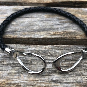 May include: A black leather bracelet with silver metal clasps. The bracelet is braided and has a simple design.