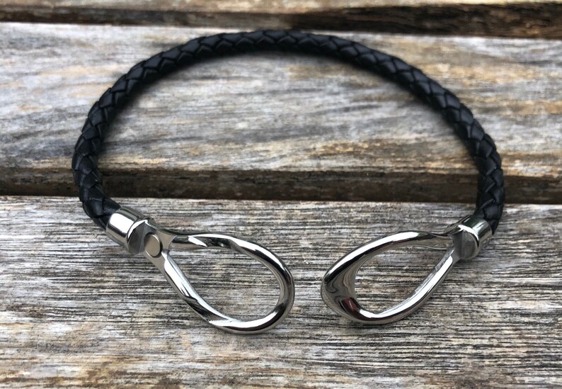 May include: A black leather bracelet with silver metal clasps. The bracelet is braided and has a simple design.