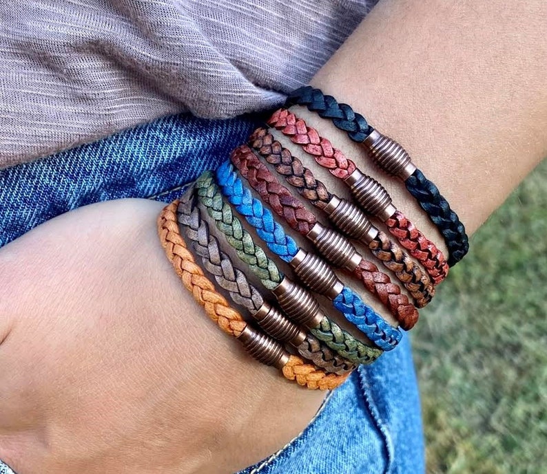 K&ouml;nnte beinhalten: Eine Auswahl geflochtener Lederarmb&auml;nder in verschiedenen Farben, darunter Orange, Grau, Gr&uuml;n, Blau, Braun und Schwarz. Jedes Armband hat einen kupferfarbenen Magnetverschluss. Die Armb&auml;nder sind an einem Handgelenk zu sehen.
