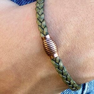 K&ouml;nnte beinhalten: Geflochtenes gr&uuml;nes Lederarmband mit kupferfarbenem Magnetverschluss. Das Armband besteht aus gewebtem, strukturiertem Leder und verf&uuml;gt &uuml;ber einen zylindrischen Verschluss mit horizontalen Rillen. Das Armband wird an einem Handgelenk gezeigt.
