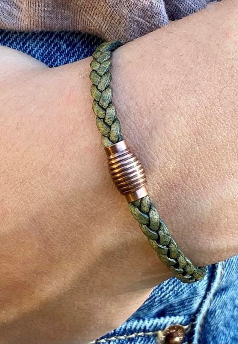 K&ouml;nnte beinhalten: Geflochtenes gr&uuml;nes Lederarmband mit kupferfarbenem Magnetverschluss. Das Armband besteht aus gewebtem, strukturiertem Leder und verf&uuml;gt &uuml;ber einen zylindrischen Verschluss mit horizontalen Rillen. Das Armband wird an einem Handgelenk gezeigt.
