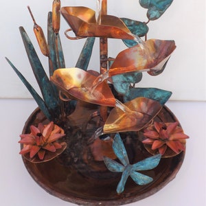 May include: A copper tabletop fountain with a cascading water feature. The fountain features a dragonfly, lotus flowers, and cattail reeds. The base of the fountain is a copper bowl.