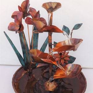 May include: A copper water fountain featuring a tiered design with cascading leaf-shaped bowls. The structure includes metallic flower accents with blue-green patina finishes, a central stem, and a circular basin filled with water at the base.