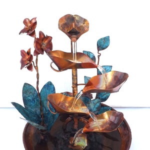 May include: A copper tabletop fountain with a cascading water feature. The fountain is decorated with copper leaves and flowers, and has a circular base.