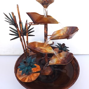 May include: A copper tabletop fountain with a cascading water feature. The fountain features a series of copper leaves and a flower at the top. The base of the fountain is a copper bowl with a water lily design.