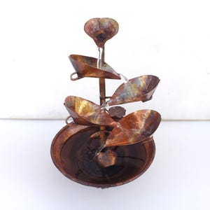 May include: A copper-toned metal fountain with cascading water flowing down a series of leaf-shaped bowls. The fountain has a rustic, aged look and is a decorative piece for a garden or home.