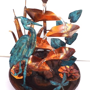 May include: A tiered copper water fountain featuring a verdigris heron standing in a basin. The fountain includes sculpted copper leaves, cattails, and two dragonfly figures. Water flows from the top leaf down through the levels into the base.