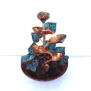 May include: A tiered water fountain featuring polished copper leaf-shaped basins and oxidised teal-green metal foliage. Water flows from the top centre down through the cascading copper leaves into a wide, circular copper basin at the base.