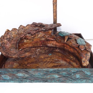 May include: A copper tabletop fountain with a rustic, wood-like design. The fountain features a small pool with a waterfall cascading over a rock formation. Two copper turtles are perched on the rock formation. The base of the fountain is decorated with a wave pattern.