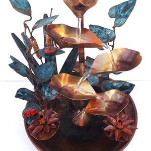 May include: A copper tabletop fountain with a cascading water feature. The fountain is decorated with copper leaves, flowers, and ladybugs. The water flows from a copper flower at the top of the fountain and cascades down into a copper bowl.