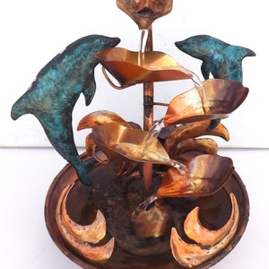 May include: A copper fountain with a cascading water feature. The fountain is decorated with two dolphins and leaves. The water flows from the top of the fountain down through the leaves and into a bowl at the bottom.