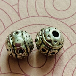 May include: Two silver metal beads with a textured, circular design. The beads have a hole in the center for stringing.