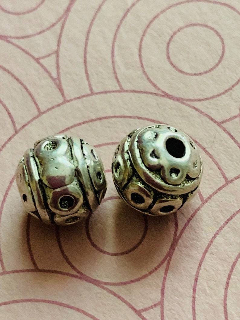 May include: Two silver metal beads with a textured, circular design. The beads have a hole in the center for stringing.