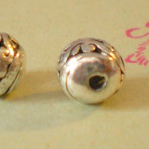 May include: Two silver beads with intricate designs. The beads are round and have a hole in the center.