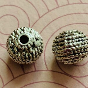 May include: Two silver metal beads with a textured surface. The beads have a hole in the center for stringing.