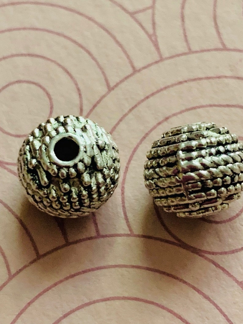 May include: Two silver metal beads with a textured surface. The beads have a hole in the center for stringing.