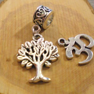 May include: Silver-coloured metal charms and beads. The image shows a tree of life charm, an Om symbol charm, and a bead with a decorative heart design. These charms are likely for jewellery making or crafting.