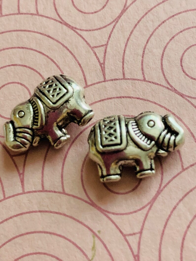May include: Two silver elephant charms with intricate details. The elephants are facing to the right and have raised trunks.