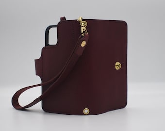 May include: A dark burgundy leather phone case with a black inner frame, featuring a gold-toned metal snap closure, a matching wrist strap attached with a gold-toned swivel clasp, and a reinforced metal eyelet at the bottom edge.