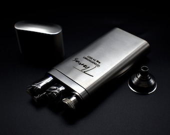 May include: A silver metal flask with a funnel and a small silver box. The flask is engraved with the text "Groomsten 22.12.2023" and "Thomas".