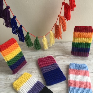 May include: Five knitted phone cases featuring different pride flag colours. The cases are arranged on a white wooden surface. A rainbow tassel garland hangs above the cases.