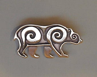 Puede incluir: Un broche de plata con forma de oso con un patrón de espiral. El oso está mirando hacia la derecha y tiene un diseño detallado.