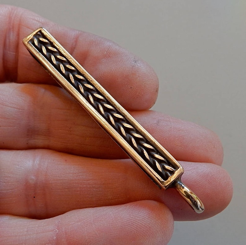 Cast bronze bar to slide onto a belt with a suspension loop at the bottom.  Herringbone wire pattern on the front in a boxed border.  By Master Ark.