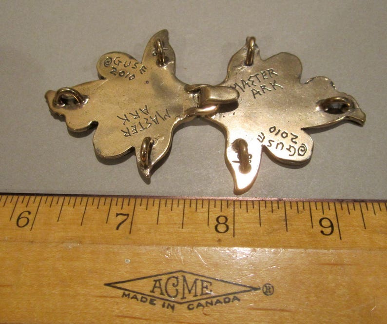 Puede incluir: Un colgante de metal dorado en forma de dos hojas estilizadas. Las hojas est&aacute;n conectadas por una barra de metal. La parte posterior del colgante est&aacute; grabada con el texto "Master Ark" y "Guse 2010".