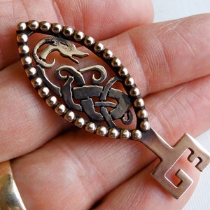 May include: A bronze key pendant with a Celtic knot design and a beaded border. The key has a stylized dragon head at the top and a simple square keyhole at the bottom.