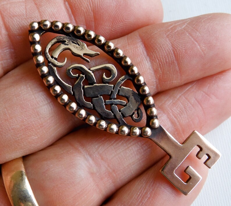 May include: A bronze key pendant with a Celtic knot design and a beaded border. The key has a stylized dragon head at the top and a simple square keyhole at the bottom.