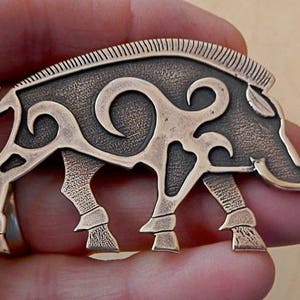 Op de afbeelding: Een bronzen everzwijn broche met ingewikkelde Keltische knoopwerk ontwerpen. De broche is gedetailleerd met een getextureerd oppervlak en een verhoogde rand langs de achterkant.