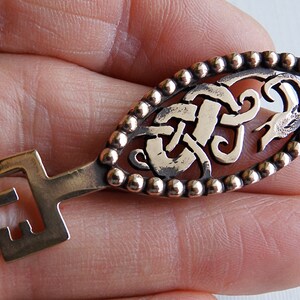 May include: A gold-toned key with a detailed Celtic knot design and a beaded border. The key has a simple, straight handle.