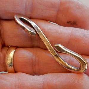 May include: A vintage, curved metal hook in a warm, brassy tone. The hook is held in a hand, with a gold ring visible on a finger. The hook's design features a looped end and a slender, curved body.