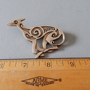 Running greyhound, in profile, facing right, with legs crossed under body.  Body is decorated with cut in Celtic spirals.  Pictured above a ruler - 3 inches long. 
 Bronze by Master Ark.