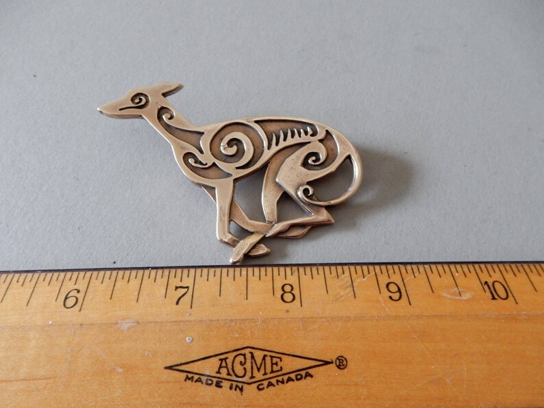 Running greyhound, in profile, facing right, with legs crossed under body.  Body is decorated with cut in Celtic spirals.  Pictured above a ruler - 3 inches long. 
 Bronze by Master Ark.