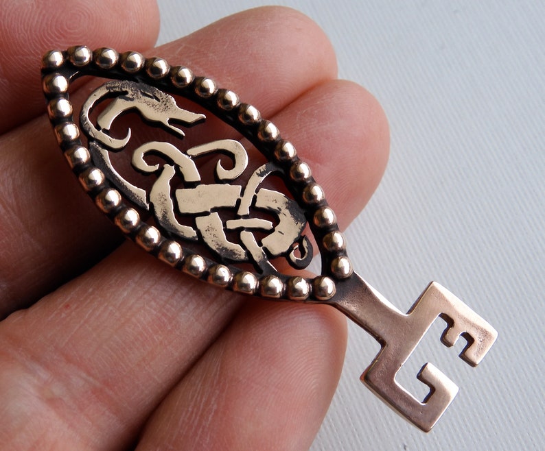 May include: A bronze key pendant with an intricate Celtic knot design and a beaded border. The key has a long, narrow head and a simple, square-shaped bow.