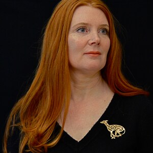 Redhead model wearing running greyhound brooch.  Greyhound is in profile, facing right, with legs crossed under body.  Body is decorated with cut in Celtic spirals.  Bronze by Master Ark.