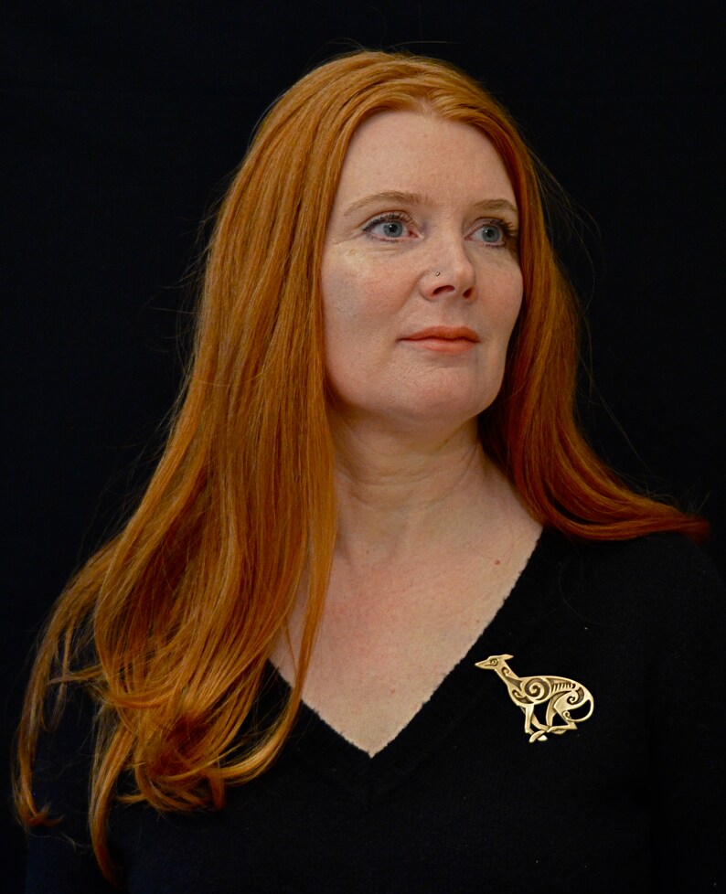 Redhead model wearing running greyhound brooch.  Greyhound is in profile, facing right, with legs crossed under body.  Body is decorated with cut in Celtic spirals.  Bronze by Master Ark.