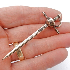 Könnte beinhalten: Eine kleine Schwertbrosche im antiken Stil mit detailliertem Griff und langer, schlanker Klinge. Die Brosche ist aus goldfarbenem Metall gefertigt und weist auf dem Griff aufwendige Designs auf. Das Schwert wird in einer Hand vor weißem Hintergrund gehalten.