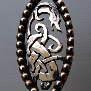 May include: A silver-colored metal spoon with a decorative handle. The handle is oval-shaped and features a Celtic knot design with a beaded border.