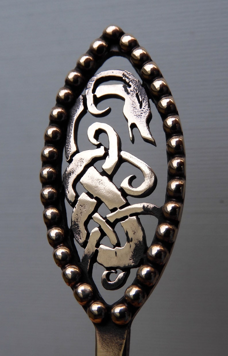 May include: A silver-colored metal spoon with a decorative handle. The handle is oval-shaped and features a Celtic knot design with a beaded border.
