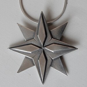 Silver coloured eight point white bronze star. Four triangular points are on the lowest layer. Four more points overlay these filling in between them.  The top four points have a raised elongated diamond shape on top of them. On chain. By Master Ark.